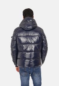 Giacca puffa navy con finitura lucida, cappuccio attaccato e motivi di cucitura orizzontale; abbinata a jeans blu.