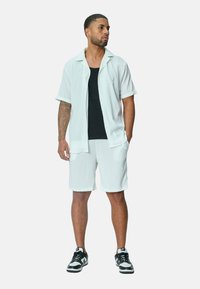 Chemise blanche à manches courtes et shorts en tissu texturé, superposés sur un débardeur noir. Des baskets noires avec des accents blancs complètent le look.