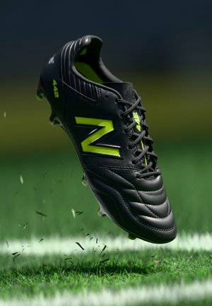 Čierny futbalový kopačka so zelenými akcentmi, textúrovaný zvršok a tvarované kolíky, zachytená vo vzduchu nad trávnatým ihriskom s mierne rozptýlenými nečistotami.