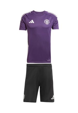 Joven con camiseta de fútbol morada del Manchester United con paneles laterales blancos y negros, de pie con las manos en las caderas.