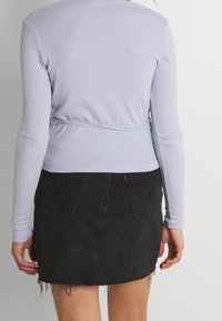 Kvinde iført en lyse-lilla langærmet bluse og en sort denim mini-skjorte med frynsede kanter, set fra bagsiden mod en hvid baggrund.