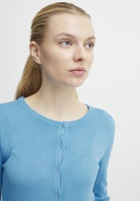 Cardigan bleu clair en matériau tricoté doux. Présente un col rond et six boutons assortis sur le devant.