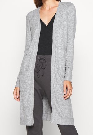 Femme portant un long cardigan gris clair ouvert sur le devant avec des poches, un haut noir à col en V et un pantalon gris foncé à cordon.