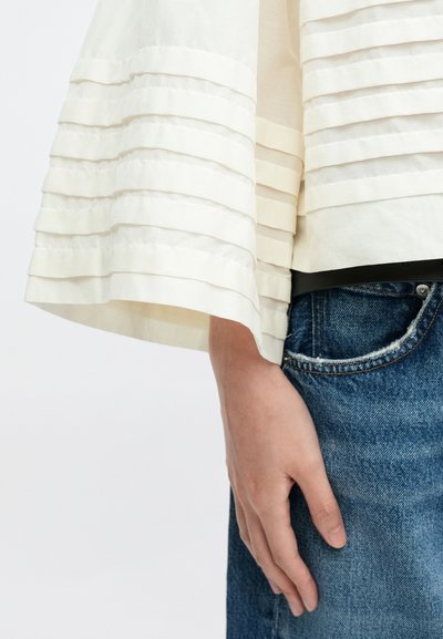 Hånd hvilende ved siden af blå denimjeans, iført en cremefarvet bluse med horisontale, plisserede lag på ærmet mod en ensfarvet baggrund.