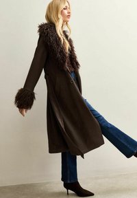 Abrigo de trench de piel sintética marrón con cuello alto, mangas largas y puños forrados de piel, combinado con vaqueros acampanados azules y tacones negros.
