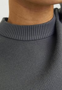Grå strikket sweater med ribbet halsudskæring, der har en struktureret overflade og blødt materiale, med nærbilleder af stoffets detaljer.