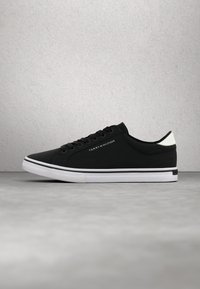 Baskets basses noires avec semelle blanche et détail au talon, lacets noirs et logo Tommy Hilfiger sur le côté, posées sur une surface grise.