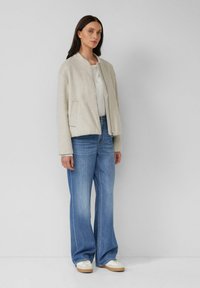 Hellgrau Bomberjacke mit Reißverschluss, seitlichen Taschen und gerippten Bündchen, kombiniert mit weit geschnittenen blauen Jeans und weißen Sneakers.