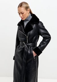 Černý kabát z umělé kůže s měkkým černým kožichem na límci a manžetách, s opaskem v oblasti pasu a zipovými kapsami. Má přiléhavý střih a dlouhé rukávy.