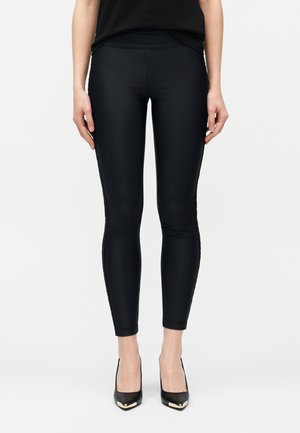 Persoană purtând colanți negri mulați și pantofi negri cu toc înalt și accente metalice aurii, stând pe un fundal simplu, deschis la culoare.