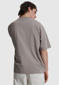 Tricou din bumbac gri, cu mâneci scurte, guler rotund și o croială lejeră, prezentând o textură netedă pe spate, expus pe un fundal deschis.