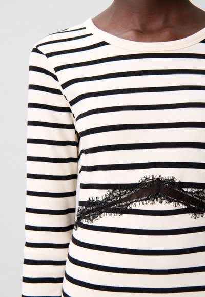 Camisa de manga larga en crema con rayas horizontales negras, con un detalle de encaje en la cintura y bordes con flecos. Material suave de algodón.