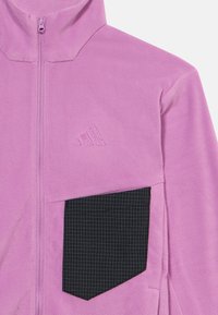 Ζακέτα φλις σε ανοιχτό ροζ χρώμα με ψηλό γιακά, πλήρες φερμουάρ και μια μεγάλη μαύρη τσέπη με σχέδιο πλέγματος στο μπροστινό μέρος.