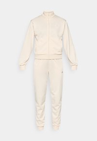 Beige träningsdräkt med hög krage, hel dragkedja och muddar. Har broderad logotyp i ljuslila på jackan och byxorna.