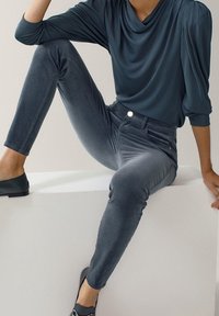 Person som sitter på en vit kub iförd en mörkblå långärmad topp med draperingar, åtsittande grå sammetbyxor och svarta slip-on skor.