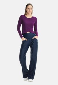 Langes lila Oberteil mit verdrehtem Saum und marineblaue, weit geschnittene Hosen mit seitlichen Taschen, hergestellt aus glattem, leichtem Stoff.