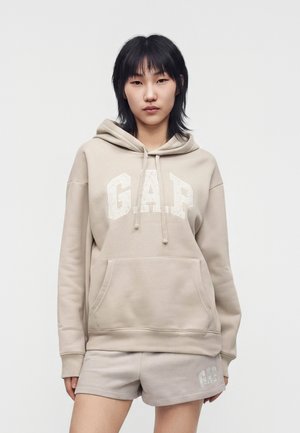 Femme aux cheveux noirs raides portant un sweat à capuche GAP beige et un short assorti, debout contre un fond clair uni, face à l'avant.