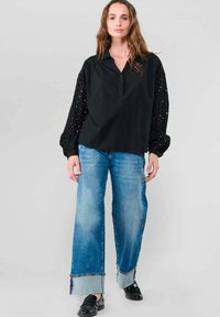 Frau in einer schwarzen Bluse mit Lochmusterärmel, weit geschnittene blaue Jeans mit weitem Bund und schwarze Slipper, die vor einem schlichten grauen Hintergrund steht.