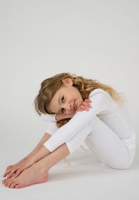 Witte lange mouwen top en bijpassende leggings, nauwsluitend, van zacht materiaal. Model poseert zittend met de armen om de gebogen knieën gewikkeld, lichte achtergrond.