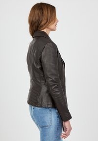 Ruskea nahkatakki, jossa on kiinteä malli, uurretut olkaaccentit ja vetoketjukiinnitykset. Yhdistetty sinisiin farkkuihin. Sivuprofiili.