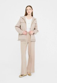 Beige jakke med to forlommer og gulddetaljer, kombineret med en hvid skjorte og matchende vidde bukser med brede ben. Glat stoffinish.