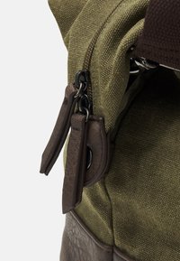 Bolso de lona en verde oliva con detalles en cuero marrón, que presenta un cierre de cremallera y tiradores de cremallera de cuero. Tejido texturizado, diseño duradero.