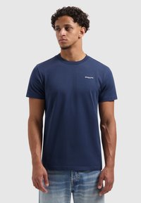 Donkerblauwe T-shirt met korte mouwen van katoen; heeft een ronde hals en een wit logo op de borst. Draagt met lichtblauwe spijkerbroek.