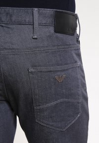 Mörkgrå denimjeans med en bakficka med en sydd design och en svart läderetikett med ett präglat logotyp.