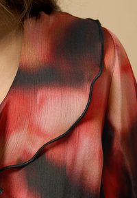 Nærbillede af rød, sort og ferskenfarvet gradient stof med en flæset krave og sort kant på en persons skulder og nakke.