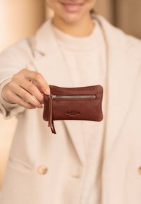 Hnědá kožená peněženka na mince se zipovým uzavíráním, která má malou koženou třásničku a embosované logo na jedné straně. Kompaktní a obdélníkový tvar.