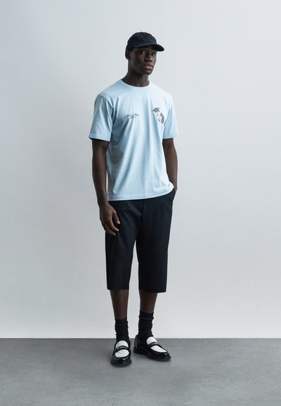 Άνδρας που φοράει ανοιχτό μπλε μπλουζάκι με σχέδιο, μαύρο σορτς μέχρι το γόνατο, μαύρο καπέλο, μαύρες κάλτσες και μαύρα με άσπρα loafers, στέκεται σε γκρι πάτωμα.