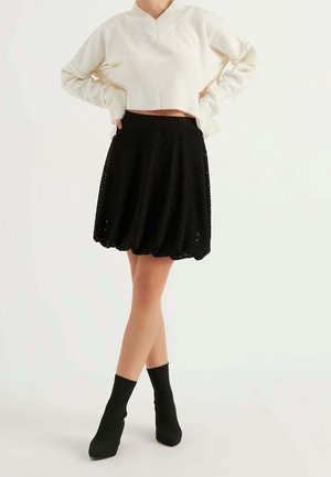 Kvinde iført en hvid cropped sweater og en sort tekstureret knælængde nederdel med sorte ankelstøvler, stående med hænderne på hofterne.