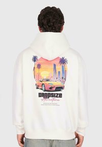 Mand iført hvid hoodie med farverig grafik af en sportsvogn, palmer og byskyer ved solnedgang med teksten "Dropsize Adventure" på ryggen.