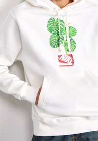 Vit hoodie med en frontficka, prydd med gröna bladjordar och en röd Nike-logga på en vit bakgrund. Snoddar och ribbade ärmslut.
