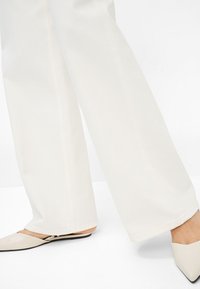 Pantalons larges en tissu blanc cassé, avec des lignes épurées et une texture lisse. Assortis à des chaussures slip-on beige clair à bout pointu.