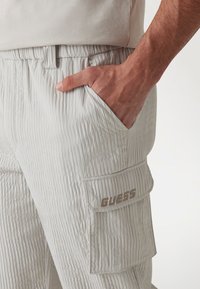 Ljusgrå ribbade cargobyxor med sidoficka; "GUESS"-logotyp i brunt på den nedre fickan. Elastisk midja och texturerad tygdetalj.