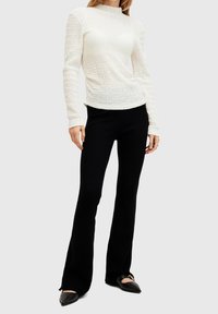 Valkoinen, teksturoitu pitkähihainen paita korkealla kauluksessa, yhdistettynä istuviin mustiin housuihin. Kengät ovat teräväkärkiset ballerinat, joissa on minimaalinen yksityiskohtaisuus.