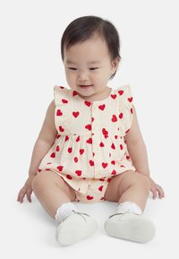 Παιδί που κάθεται και χαμογελά, φοράει εκρού ολόσωμο φορμάκι με κόκκινη καρδιά, λευκές κάλτσες και μαλακά λευκά παπούτσια σε απλό λευκό φόντο.