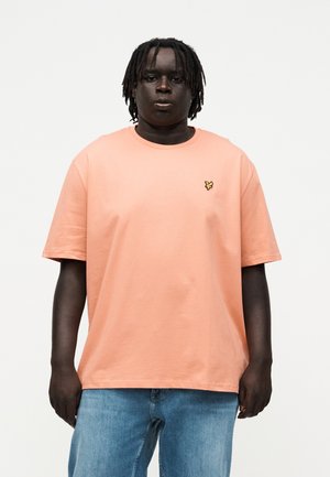 Mężczyzna o ciemnej karnacji i dredach, ubrany w brzoskwiniowy t-shirt z krótkim rękawem z małym żółtym logo oraz jasnoniebieskie dżinsy, stojący na jednolitym białym tle.