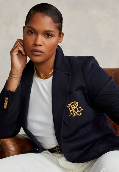 Femme aux cheveux courts portant des boucles d'oreilles créoles en or, un collier chaîne en or, un blazer bleu marine avec un logo brodé en or et un haut blanc, assise sur une chaise marron.