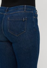 Jean en denim bleu foncé avec une coupe ajustée. Comprend une poche arrière et des détails de couture. Texture lisse avec un minimal de distorsion.
