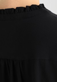 Blusa nera in tessuto texturizzato con un colletto alto arricciato e dettagli arricciati sotto lo yoke.