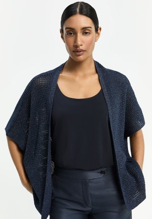 Frau mit dunklen Haaren in einem tiefen Dutt, trägt ein ärmelloses Top in Marineblau, eine offene Strickjacke mit kurzen Ärmeln in Marineblau und marineblaue Hosen, steht mit den Händen in den Taschen.