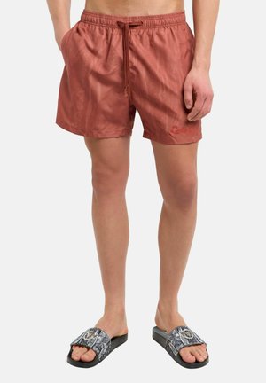 Mann trägt rostfarbene Badeshorts mit Kordelzug und gemusterte schwarze Slides, steht vor weißem Hintergrund.