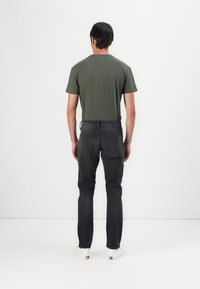 Mees, kes kannab tumehalli teksapükse, oliivrohelist t-särki ja valgeid tossusid, seisab seljaga valge seina poole.