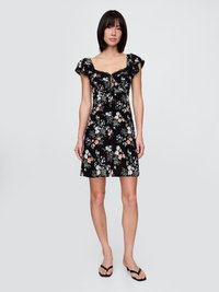Vrouw in een zwart bloemen mini-jurk met ruche mouwen en vierkante halslijn, gecombineerd met zwarte open-teven sandalen, staande op een lichte achtergrond.