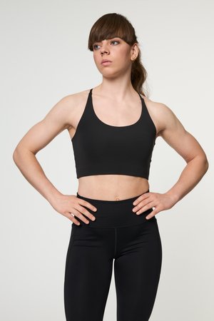 Barna hajú nő fekete sportmelltartóban és leggingsben, kezét csípőjén tartva áll, enyhén oldalra néz egy egyszínű háttér előtt.