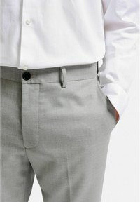 Ljusgrå, skräddarsydda byxor gjorda av texturerad tyg, med platt front, slim fit och diskreta sidfickor. Svart knappdetalj synlig.
