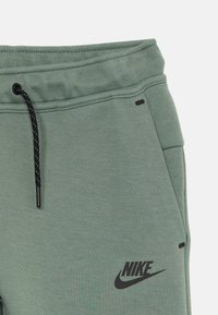 Zaļie kokvilnas maisījuma sporta bikses ar ribotu jostu, melnu auklu, sānu kabatām un melnu Nike logotipu priekšpusē.