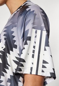 Αθλητική κοντομάνικη μπλούζα σε γκρι και άσπρο με γεωμετρικό μοτίβο· περιλαμβάνει τρεις λευκές ρίγ ες στο αριστερό μανίκι.